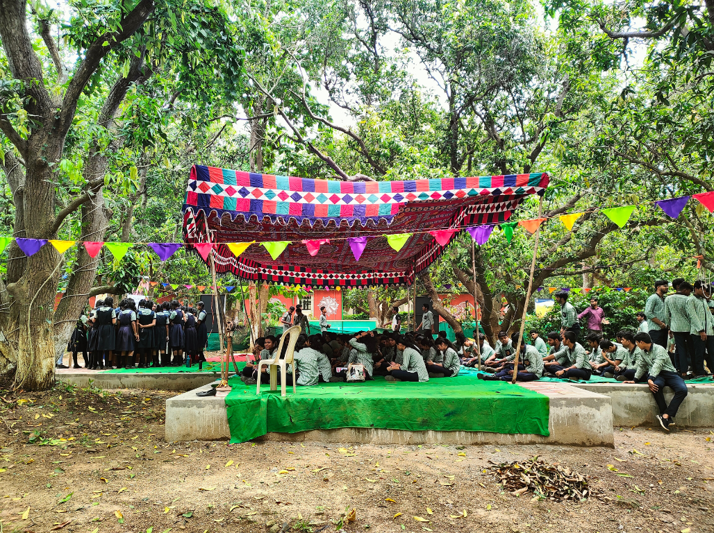 School students seated under a colourful canopy in a grove of trees during a forest camp