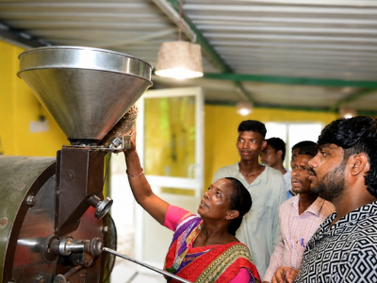 A woman feeding coffee beans into a roasting machine while young trainees watch