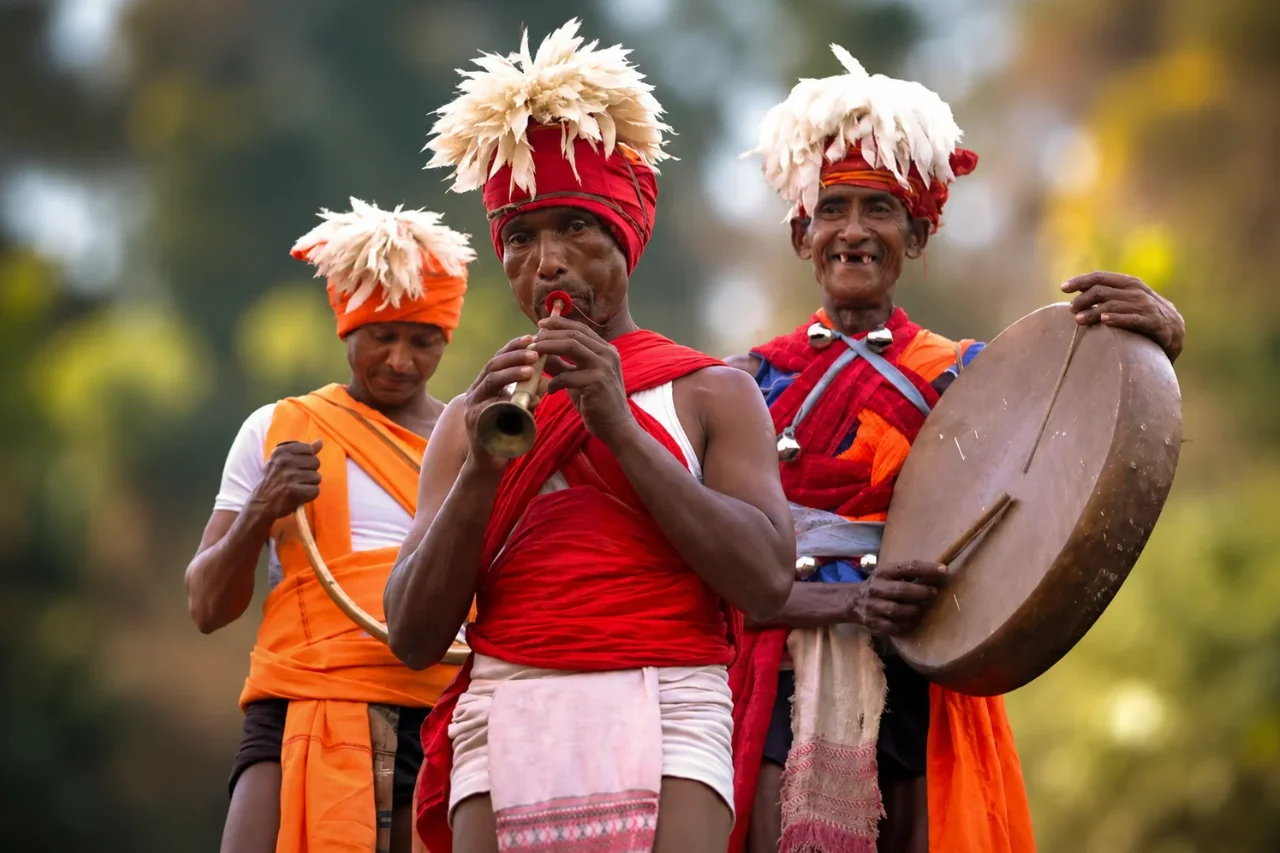 Tribal musicians in traditional dress perform with flute, drum and horn in an outdoor village setting.