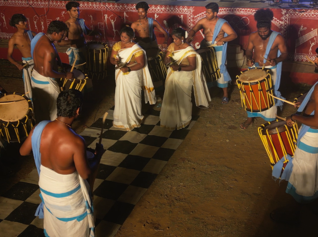 A circle of drummers in white dhotis performing before a painted mural