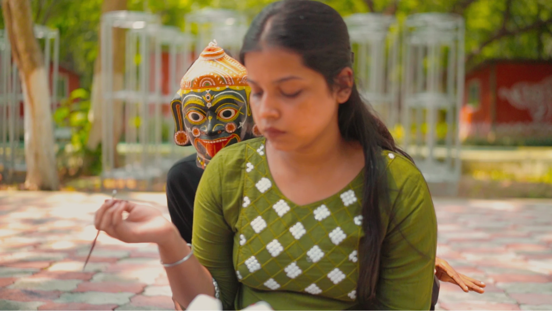 A young woman painting beside a hand-painted Chhau mask in a shaded courtyard