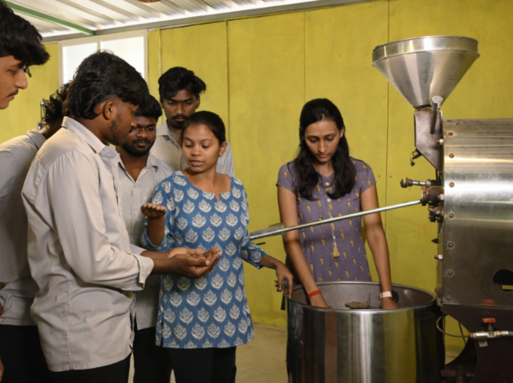 Trainees examining roasted coffee beans beside a working drum roaster