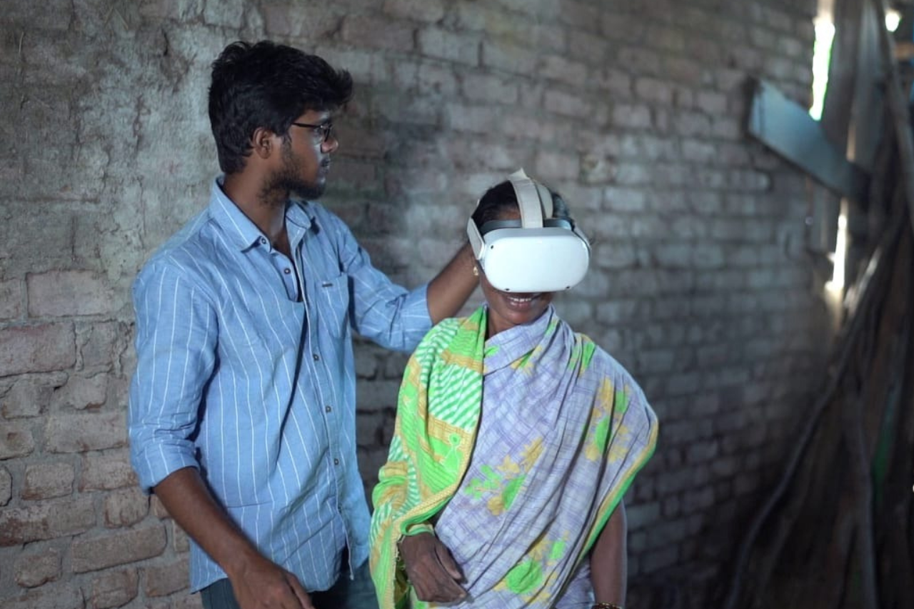 services-01 Smiling tribal woman adorned in vibrant headdress and beads against a terracotta wall in Odisha.